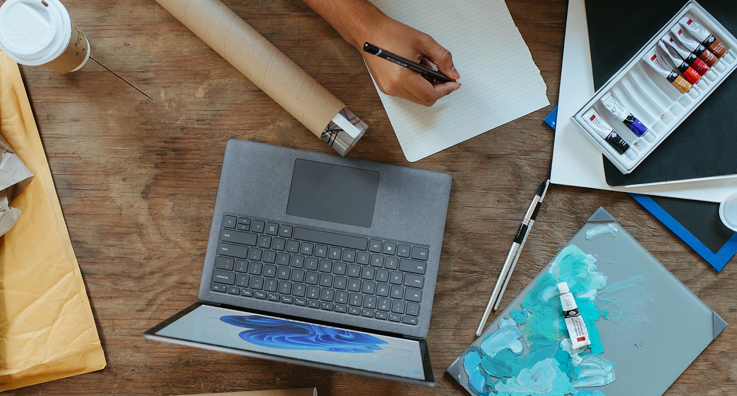 A laptop sitting on a wooden desk next to an art notebook with someone drawing in it