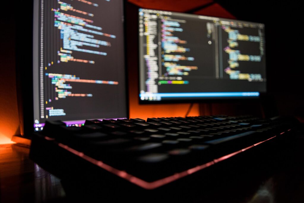 two computer monitors displaying code with a keyboard in the foreground all sitting in a dark room with orange lighting in the background